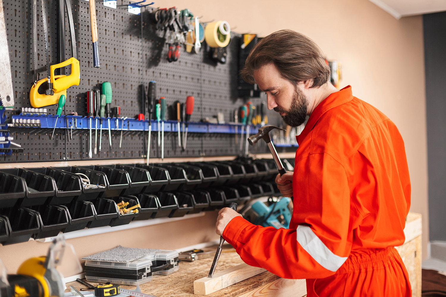 Builder in orange work clothes using hammer with stand of different tools near in workshop